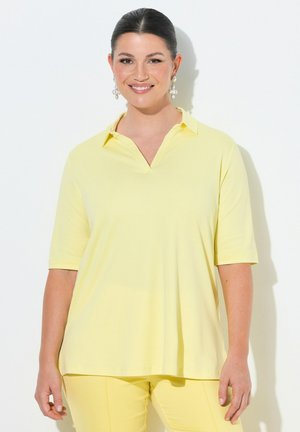 Kobieta o ciemnych włosach, nosząca perłowe kolczyki, jasnożółtą koszulkę polo z krótkim rękawem i pasujące żółte spodnie, uśmiechająca się na białym tle.