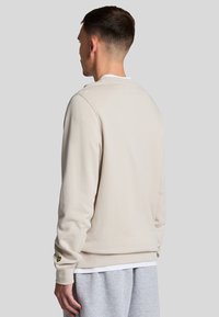 Sudadera de color beige con puños y dobladillo acanalados. Presenta un pequeño logo bordado en la manga izquierda. Textura suave, ajuste relajado.