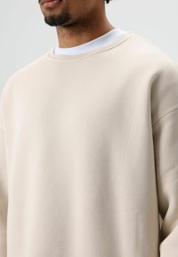 Sweat-shirt beige en coton avec col rond, coupe décontractée et épaules tombantes. Tissu légèrement texturé avec une finition lisse.