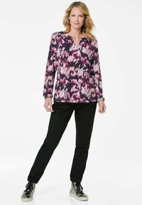 Blumenbluse mit langen Ärmeln in Lila- und Schwarztönen; kombiniert mit schwarzen Hosen und metallisch silbernen Turnschuhen.
