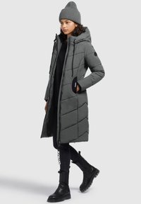 Donna che indossa un lungo cappotto grigio trapuntato con cappuccio, pantaloni neri, stivali neri e un berretto grigio in maglia, in piedi su uno sfondo neutro.