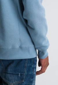 Lichtblauwe sweatshirt met ribgebreide boorden en onderkant, gecombineerd met middelblauwe denim jeans met een klein wit logo op de achterzak.