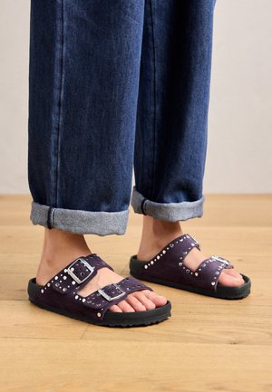 Fioletowe sandały zamszowe z dwoma regulowanymi klamrami, srebrnymi ćwiekami na paskach i czarną gumową podeszwą; stylizowane z podwiniętymi ciemnymi dżinsami.