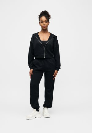 Kvinde iført sort hættetrøje med lynlås, matchende joggingbukser, hvide sneakers og minimal smykker står foran en ensfarvet hvid baggrund.