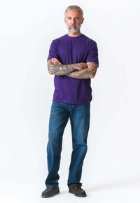 Mand i middelalderen med grå skæg og tatoveringer på armene står med armene korslagte, iført en lilla t-shirt, blå jeans og brune sko.