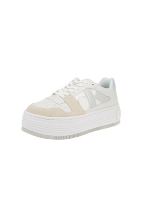 Białe sneakersy z zamszowym i skórzanym wykończeniem, z grubą gumową podeszwą, okrągłym noskiem i wyraźnym logo po boku. Pionowe żebrowane detale na podeszwie.