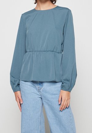 Femme portant un chemisier bleu à manches longues avec taille froncée, associé à un jean taille haute bleu clair, debout avec les bras détendus.