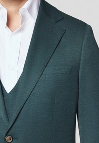 Zbliżenie osoby w ciemnozielonej, teksturowanej marynarce na białej koszuli zapinanej na guziki.