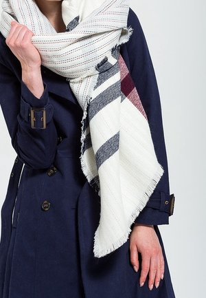 Personne portant un trench-coat bleu marine et tenant une grande écharpe blanche à rayures bleu marine et bordeaux enroulée autour des épaules.