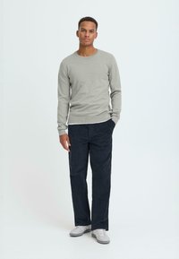 Un pulover tricotat gri deschis cu guler rotund, asortat cu o pereche de pantaloni din catifea albastră închisă și adidași albi, stând în fața unui fundal simplu.