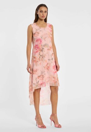 Frau steht in einem ärmellosen, rosa Blumenkleid mit asymmetrischem Saum, gerüschtem Ausschnitt und pinken High-Heel-Sandalen mit Knöchelriemen vor weißem Hintergrund.