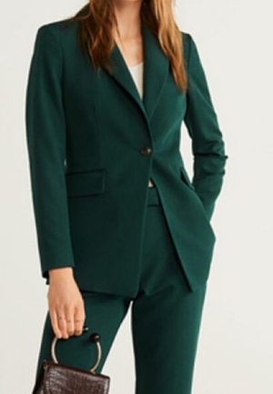 Femme portant un costume vert foncé sur mesure tenant un petit sac à main marron avec une poignée ronde dorée.
