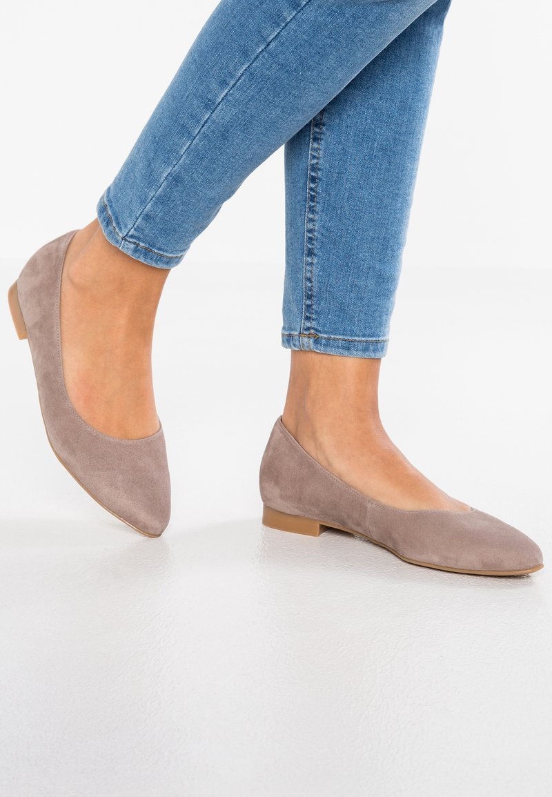 Des pieds chaussés de ballerines en daim marron clair associées à un jean slim bleu, posés sur une surface blanche et lisse.