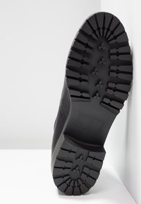 Musta saappaanpohja, jossa syvä kuviointi. Valmistettu kumista, ja siinä on suuret urat sekä pienet pitojulkuset tarttumista ja kestävyyttä varten.