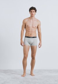 Szare bawełniane bokserki z czarnym elastycznym pasem w talii z logo. Model stoi boso na jasnoszarej powierzchni.