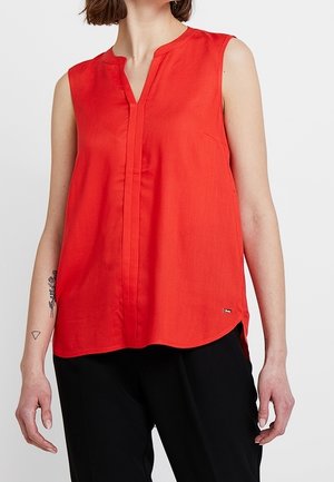 Femme portant un haut rouge sans manches à col en V et un pantalon noir, montrant des tatouages sur l'avant-bras gauche.