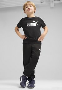 Juodas medvilninis marškinėliai su balta "PUMA" logotipu, derinami su juodomis sportinėmis kelnėmis su mažesniu logotipu. Dėvimi su tamsiai mėlynais sportbačiais.