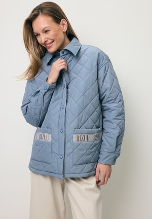 Kobieta uśmiechająca się, ubrana w jasnoniebieską pikowaną kurtkę z przednimi kieszeniami oznaczonymi napisem "BIZUU" oraz beżowe spodnie na jednolitym tle.