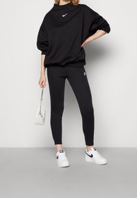 Black oversized sweatshirt with a crew neck and white Nike logo, paired with tapered black leggings and white sneakers. Silver chain handbag.