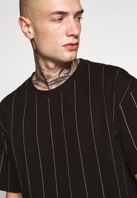 T-shirt en coton noir avec des détails de couture blanche verticale. Il présente un col rond et des manches courtes, affichant une texture lisse et mate.