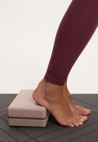 Leggings bordo cu o textură netedă, picior pe un block de yoga roz cu accente maro, așezat pe o saltea texturată neagră.