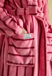 Różowy pluszowy szlafrok w czerwone paski, z paskiem do wiązania i kieszenią z etykietą "Joules HIBERNATE". Miękka, teksturowana tkanina.