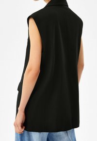 Svart ärmlös blazer med skräddarsydd design, mjuk struktur och lös passform, kombinerad med ljusa jeansbyxor.