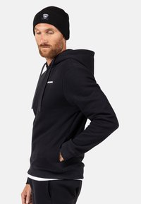 Schwarzer Hoodie mit Kängurutasche und Kapuze mit Kordelzug. Verfügt über ein weißes Logo auf der Brust und gerippte Bündchen. Kombiniert mit einer schwarzen Mütze.