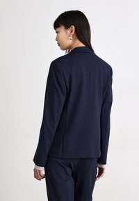 Námořnický modrý přizpůsobený blazer, směsová tkanina s vlnou, strukturovaný design, jeden rozparek na zadní straně, dlouhé rukávy, v kombinaci s odpovídajícími kalhotami.