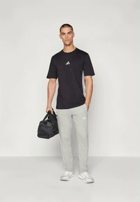 Melns sporta krekls ar baltu logo, savienots ar gaiši pelēkiem sporta biksēm ar sānu svītrām, valkāts ar baltām kedām un melnu sporta somu.