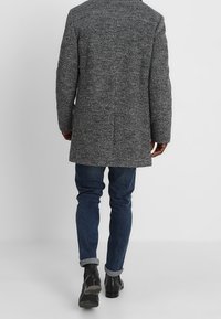 Grijze textuurjas met een getailleerde pasvorm, tot de knie, gecombineerd met blauwe jeans en zwarte enkellaarsjes. Heeft een enkele achterventilatie.