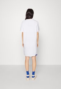 Frau, die mit dem Rücken zur Kamera steht, trägt ein weißes Kurzarmkleid, blaue Socken und beige, flauschige Sandalen vor einem schlichten Hintergrund.