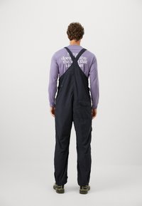 Navy blue overalls with adjustable straps, featuring a tapered leg design and a back pocket. Worn over a long-sleeve purple shirt.