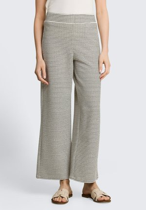 Femme portant un pantalon beige à jambes larges avec de fines rayures horizontales noires et des sandales beige, debout devant un fond uni.