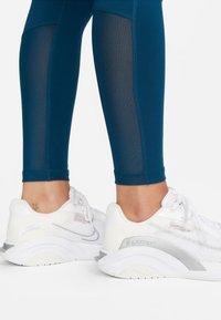 Tealfärgade träningsleggings med meshpaneler, tillsammans med vita sneakers med meshdetaljer och grå accent på sidorna och sulan.