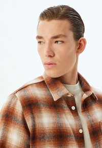 Bruin en wit geruit flanellen shirt met een kraag, voorzien van een gestructureerd patroon en knoopsluitingen. Gedragen over een beige trui.