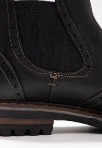 Černá kožená bota s texturovanou tkaninou, detailním prošíváním a robustní podrážkou. Má zvýrazněné perforace a tradiční design.