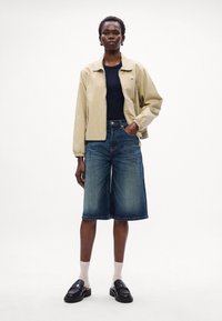 Beige bomber jaka, tumši zils tops, tumšu džinsu Bermudu šorti, melnas slīdējošas kurpes un gaišas zeķes; ikdienišķs apģērbs uz neitrāla fona.
