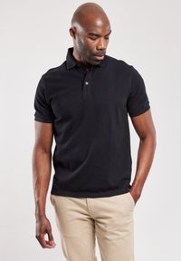 Zwarte poloshirt met korte mouwen, knoopsluiting bij de kraag en subtiele logodetails. Draagt met khaki broek, waarbij de gladde stoftextuur wordt getoond.