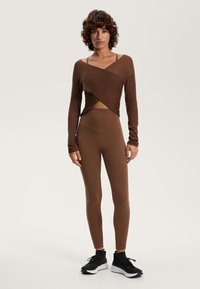 Barna bordás, hosszú ujjú felső átlós kialakítással, amelyet magas derekú barna leggingssel és fekete sportcipővel viselnek. Az anyag sima tapintású.