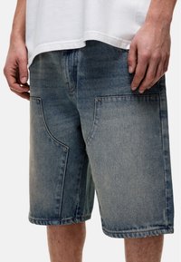 Jasnoniebieskie szorty dżinsowe z wyblakłym wyglądem. Mają duże, naszywane kieszenie, widoczne szycie oraz luźny krój.