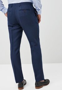 Marineblaue maßgeschneiderte Hose mit vertikalen Nadelstreifen, ausgestattet mit zwei Gesäßtaschen und einem subtilen Glanz. Kombiniert mit schwarzen Schuhen.