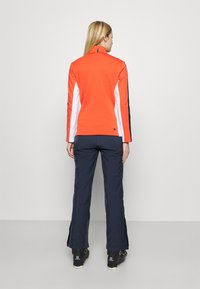 Lysoransje zip-up-genser med hvite og svarte ermer, kombinert med mørkeblå bukser. Begge laget av glatt, elastisk stoff. Svarte sko synlige.