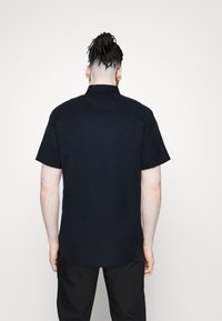 Camicia a maniche corte di colore blu navy scuro realizzata in tessuto morbido, con taglio dritto e colletto a bottone; presenta un orlo arrotondato sul retro.