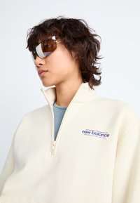 Pull en crème avec col zippé et texture côtelée. Présente un logo 'New Balance' en bleu et rouge. Le modèle porte des lunettes de soleil réfléchissantes.