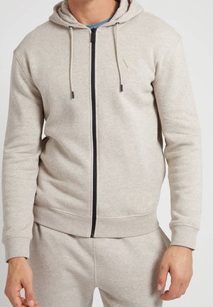 Sudadera con cremallera de color beige claro, hecha de una tela suave, con una cremallera y cordones negros, y un pequeño logo en forma de triángulo en el pecho.