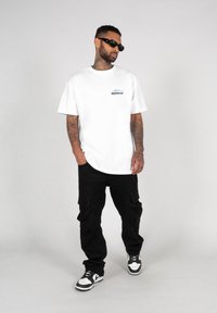 Hvit t-skjorte med en liten blå og svart logo, sammen med svarte cargo-bukser med flere lommer, og svarte og hvite joggesko.