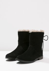 Botines de ante negros con un ribete de pelo sintético, punta redondeada y tacón bajo. Presenta un detalle decorativo de lazo en la parte trasera.