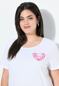 Camiseta blanca de algodón, con cuello redondo y un gráfico de un oso rosa en el lado izquierdo del pecho. Manga corta, corte relajado.