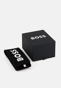 Boîte noire avec le logo "BOSS" en texte blanc, à côté d'un article en tissu noir portant le même logo. Le tissu a une texture lisse et un détail contrastant.